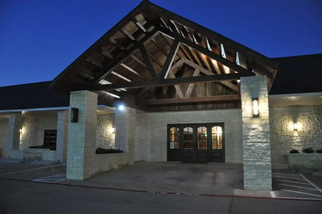 A stone building entrance at dusk featuring a large wooden gable roof and bright lighting above double glass doors.