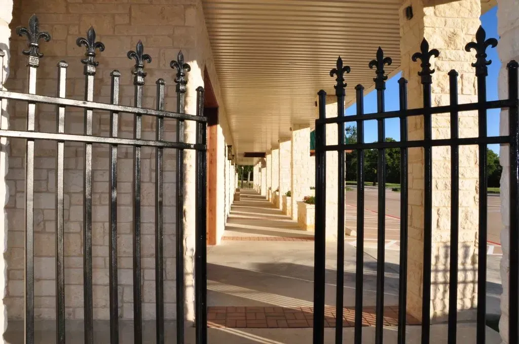 A black wrought-iron fence stands before a covered stone walkway, leading to a sunlit exterior area.