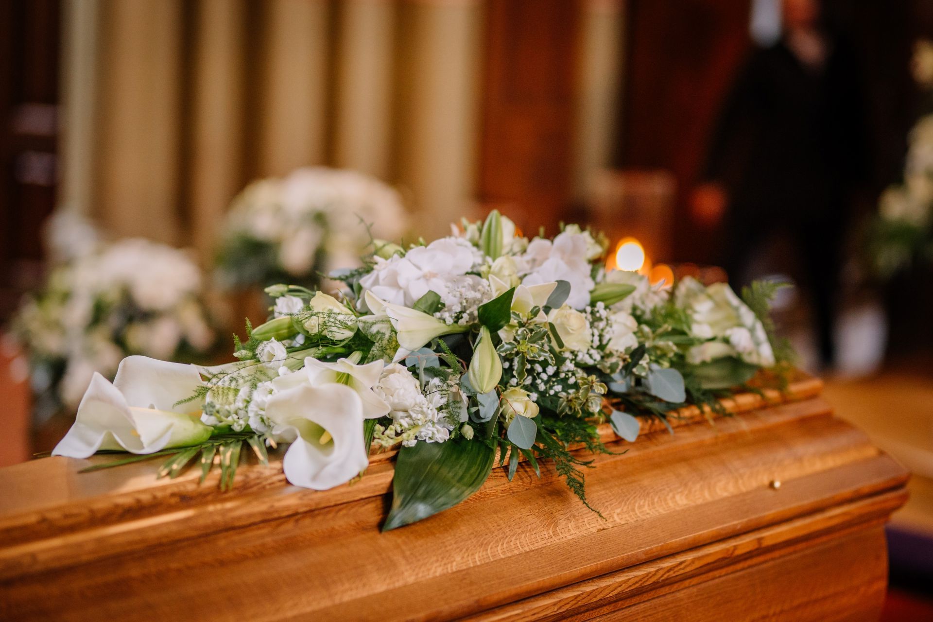 Wooden casket with white floral arrangement; church interior; blurred figure in background.