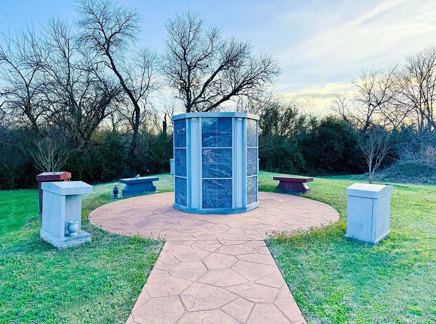 A stone columbarium stands on a circular patio, surrounded by small stone markers and trees in a cemetery at sunset.