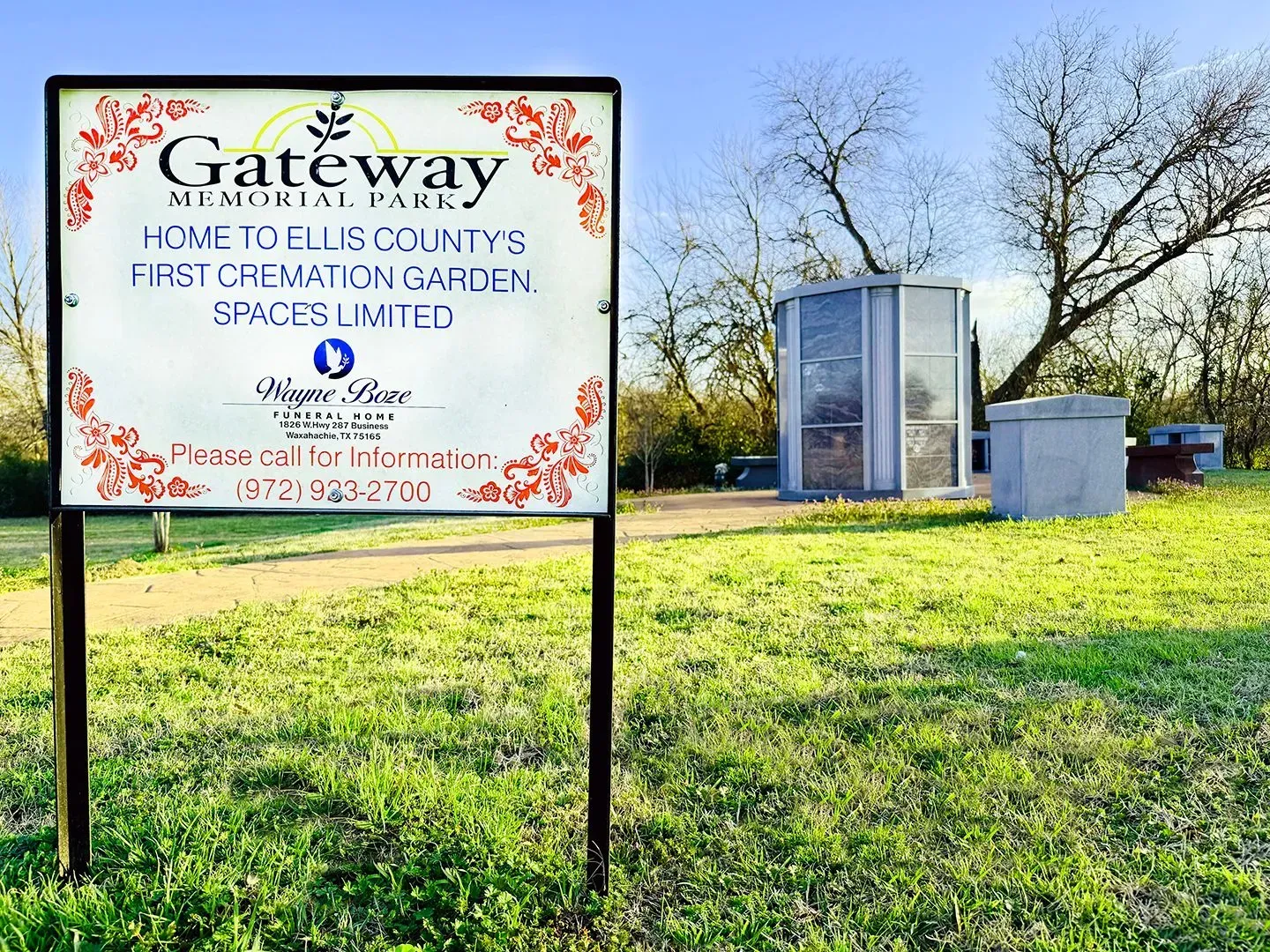 A Gateway Memorial Park sign stands in a grassy cemetery field with a cremation garden structure in the background.