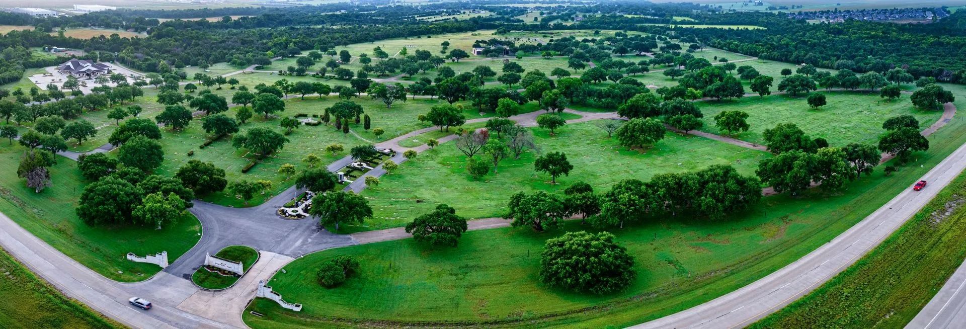 Aerial view of a green park with trees and roads. Cars are parked near the entrance.