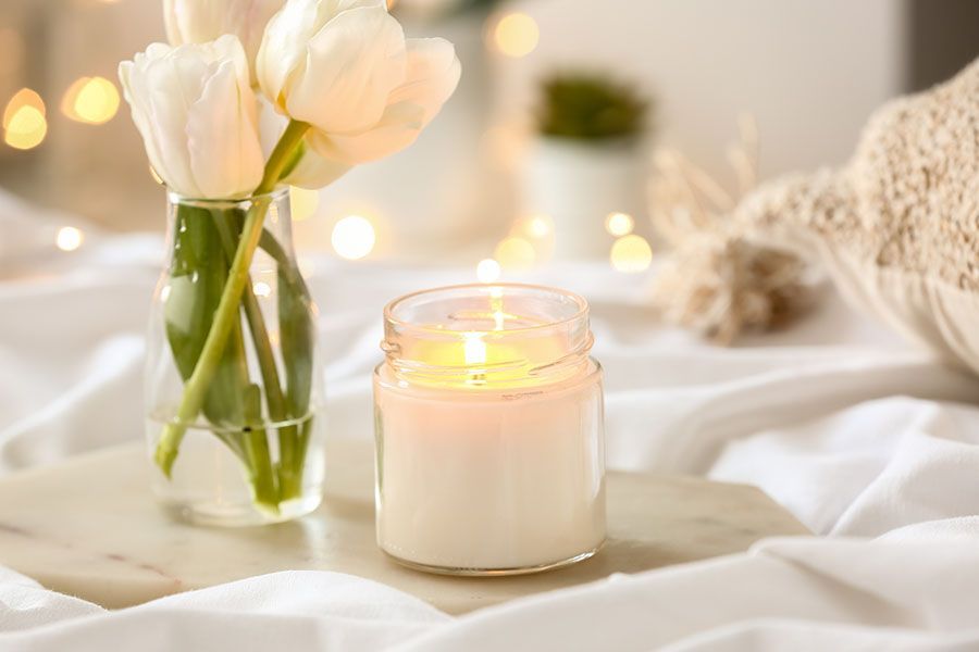 Candle burning next to white tulips in a glass vase on a white cloth, with soft lighting.