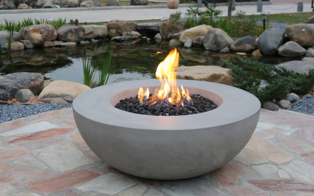 A round fire bowl with flames, placed on a stone patio near a pond