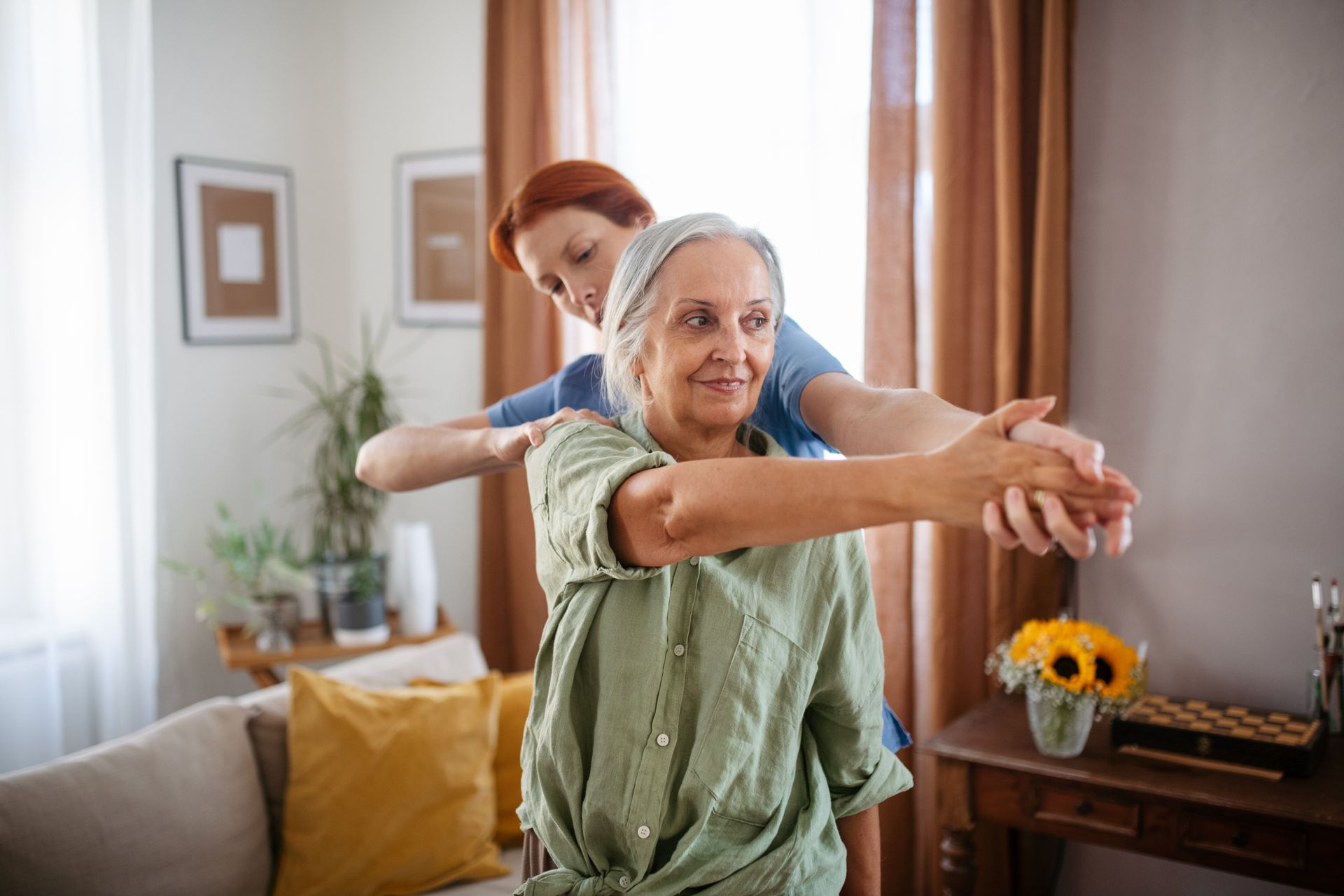 A nurse is helping an elderly woman stretch her arms in a living room.