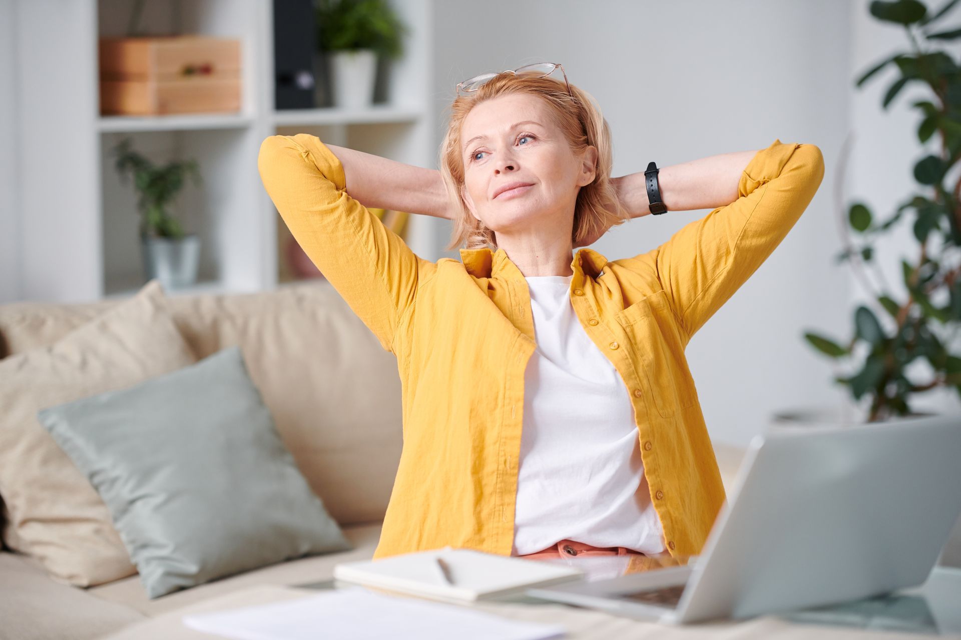 A woman is sitting on a couch with her hands behind her head in front of a laptop computer.