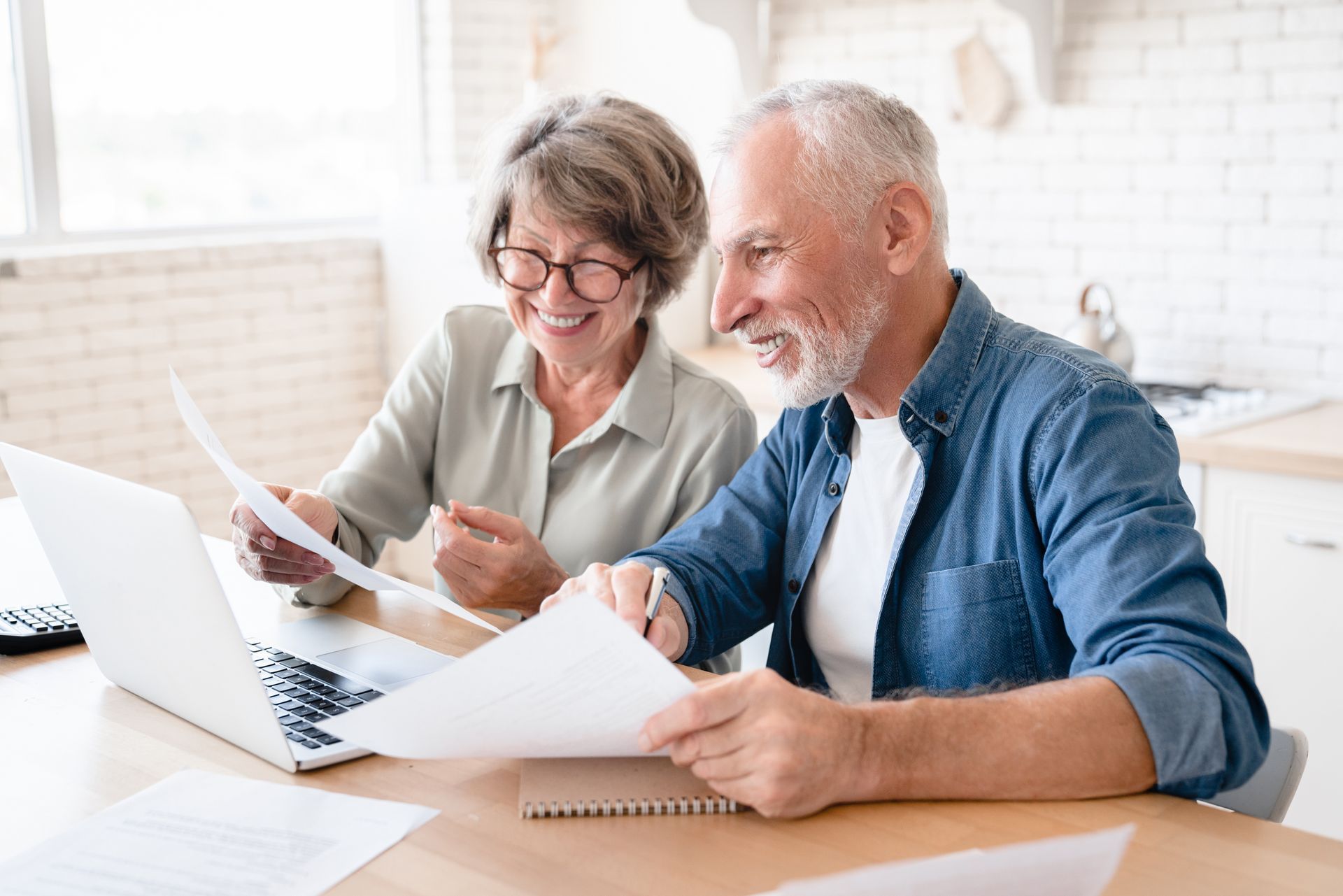 An elderly couple is sitting at a table looking at papers and a laptop.