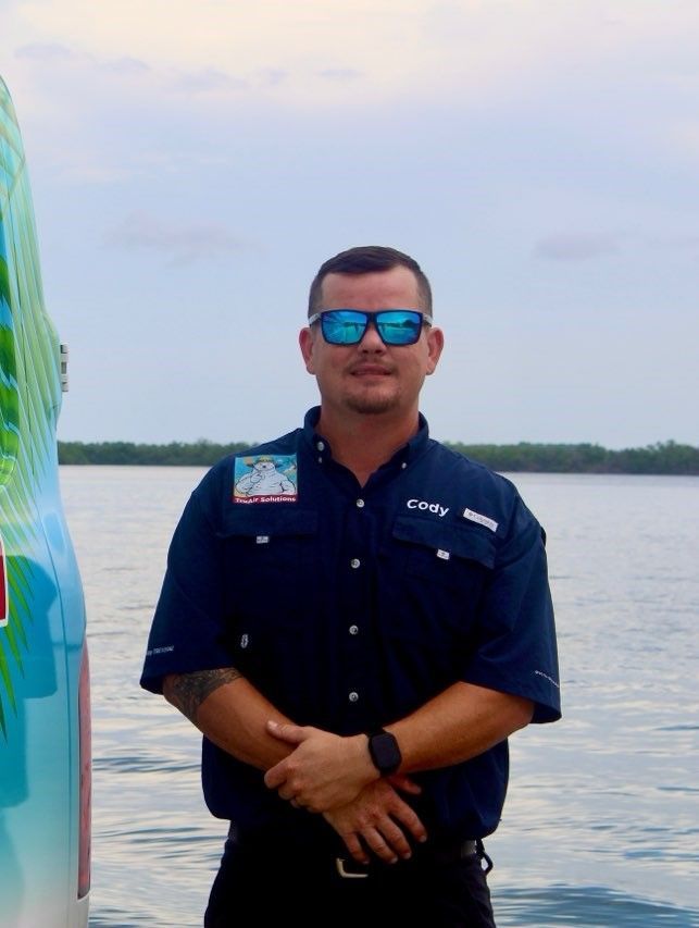 Man wearing sunglasses and a blue shirt, standing in front of a body of water with arms crossed.