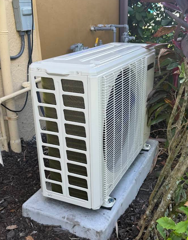 Outdoor air conditioning unit on a concrete base, near a building with vegetation.