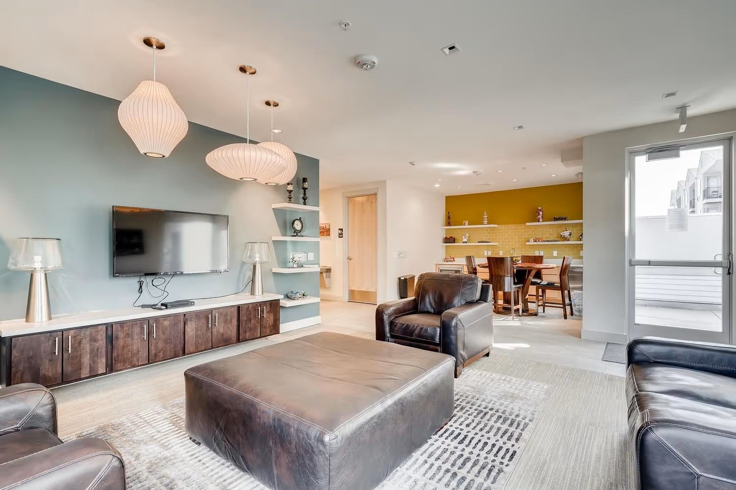 A modern living area with a large ottoman, leather armchairs, a TV on a blue accent wall, and a dining space in the back.