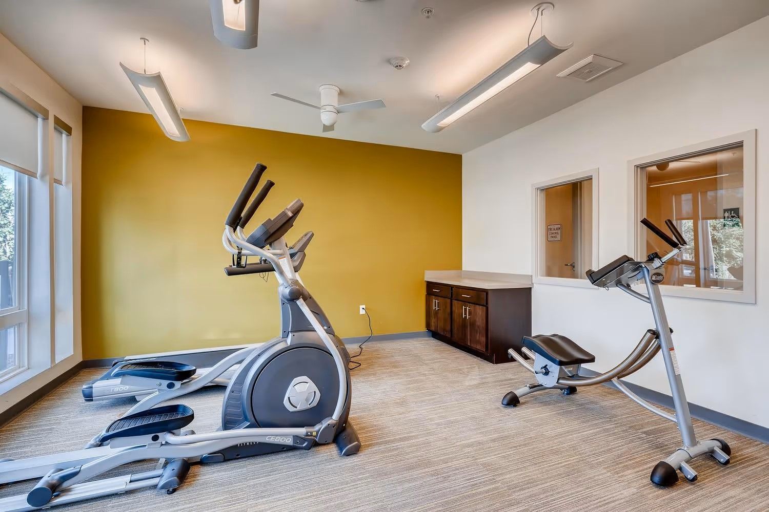A gym with an elliptical machine and a seated exercise bike, featuring a mustard-yellow accent wall and overhead lighting.