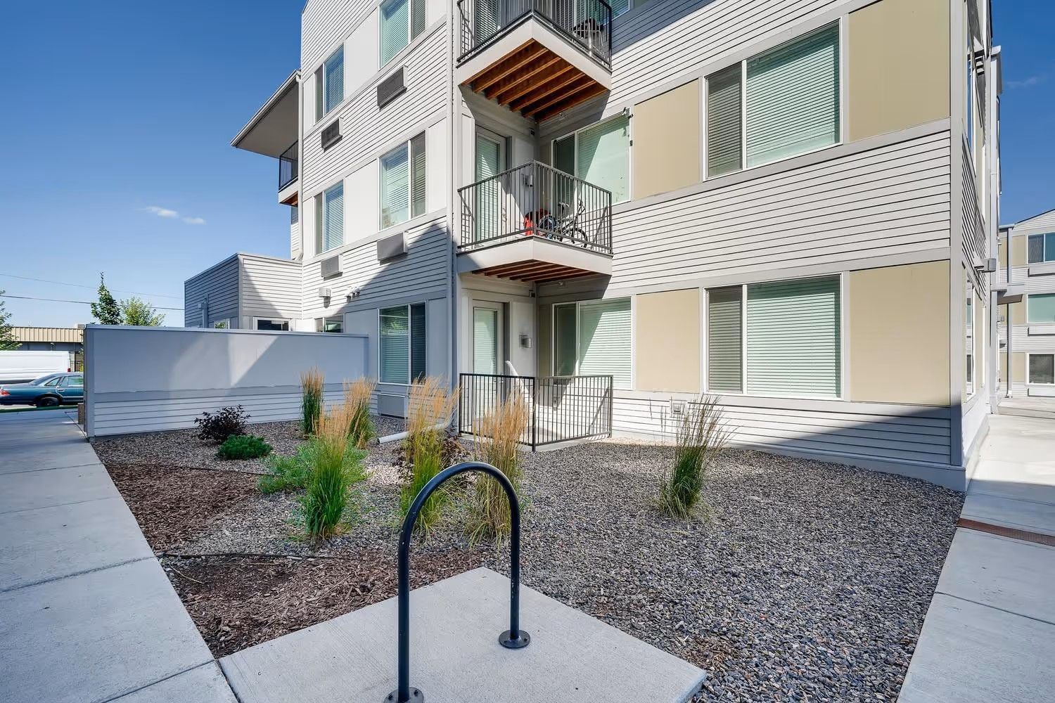A three-story apartment building with beige siding and balconies, next to a landscaped courtyard with a bike rack.