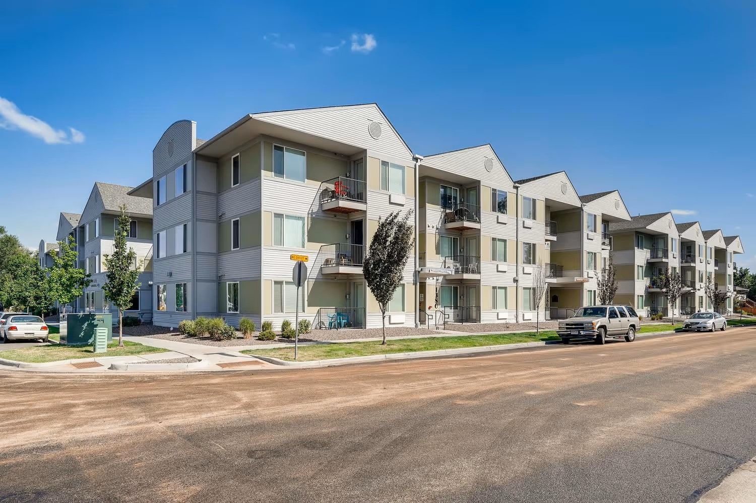 A multi-story apartment building with beige siding and balconies under a clear blue sky, viewed from a street.