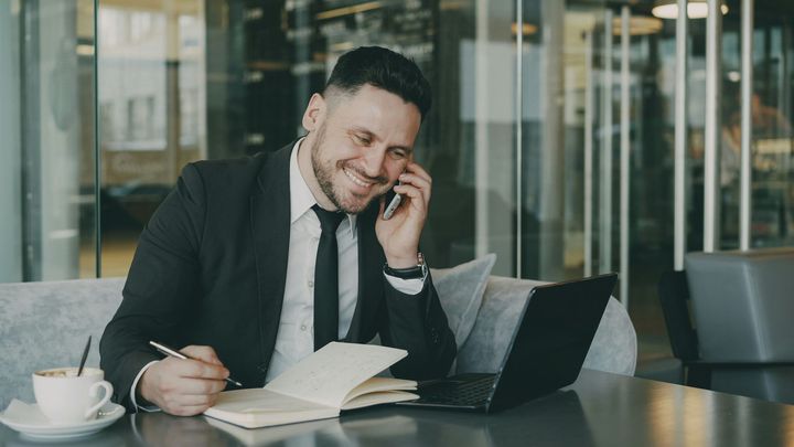 A professional in a suit and tie smiling while talking on a phone, writing in a notebook next to a laptop and coffee.