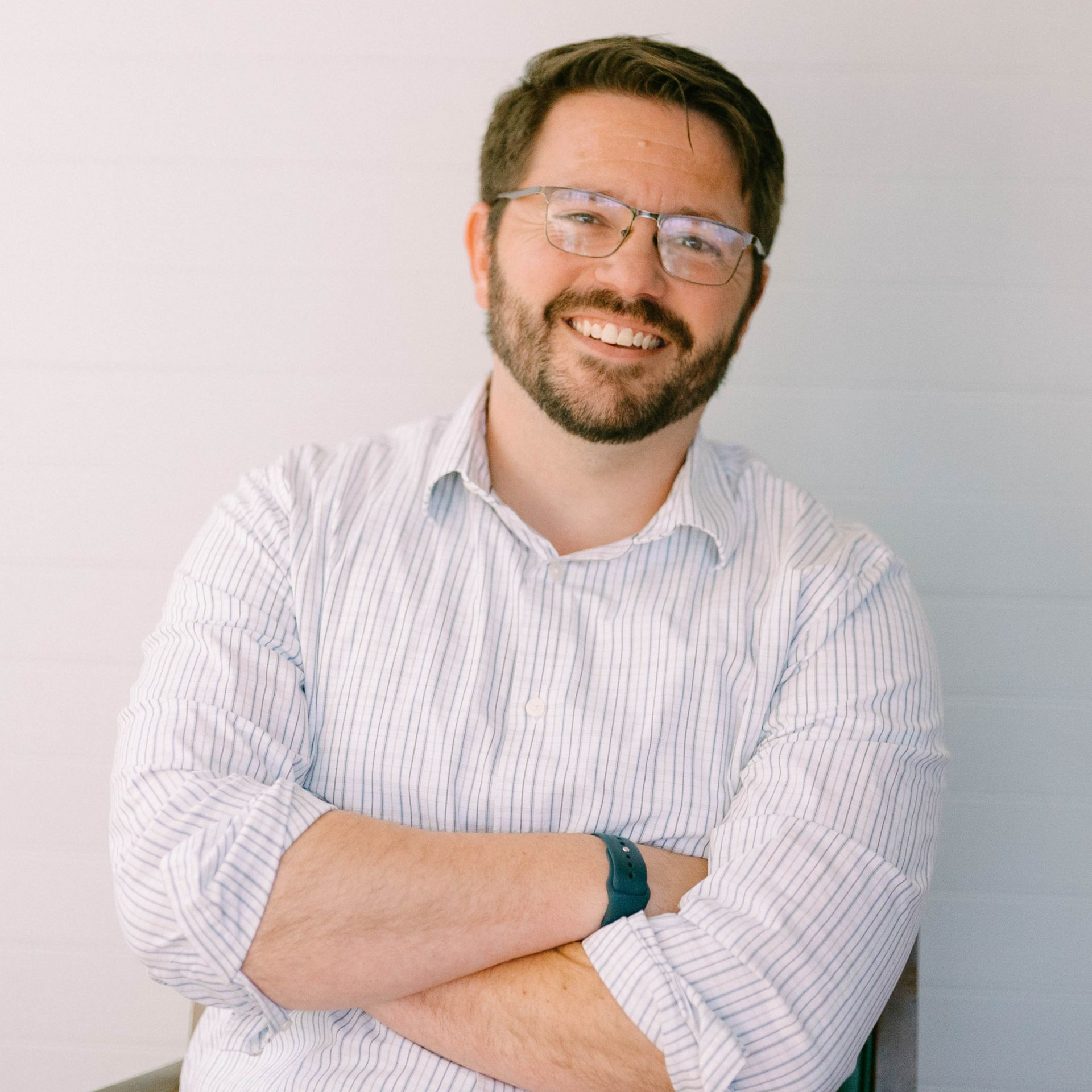 Man with arms crossed, smiling, wearing a blue button-down shirt, standing outside a building.