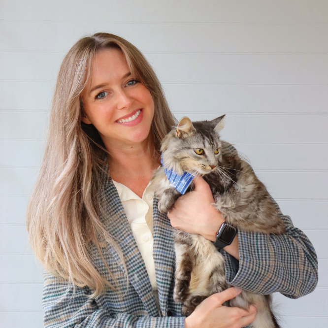 Woman with blonde hair smiles, holding a gray cat wearing a blue bow tie, standing in front of a white wall.