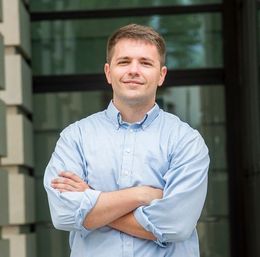 A man in a blue shirt is standing with his arms crossed in front of a building.