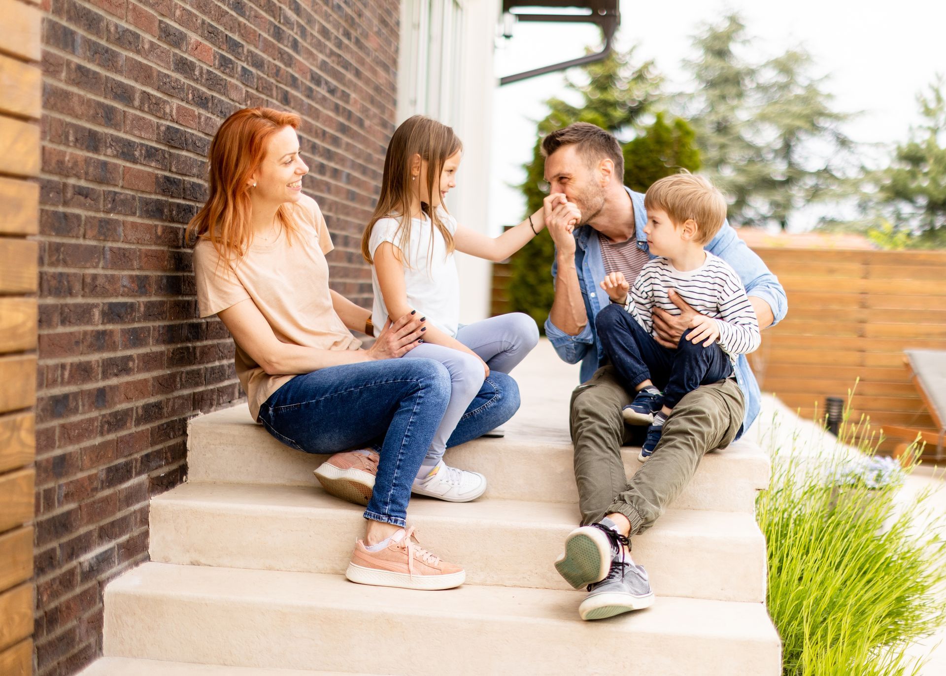 Family sitting on steps, interacting. Smiling, hugging, near brick wall and wooden fence.
