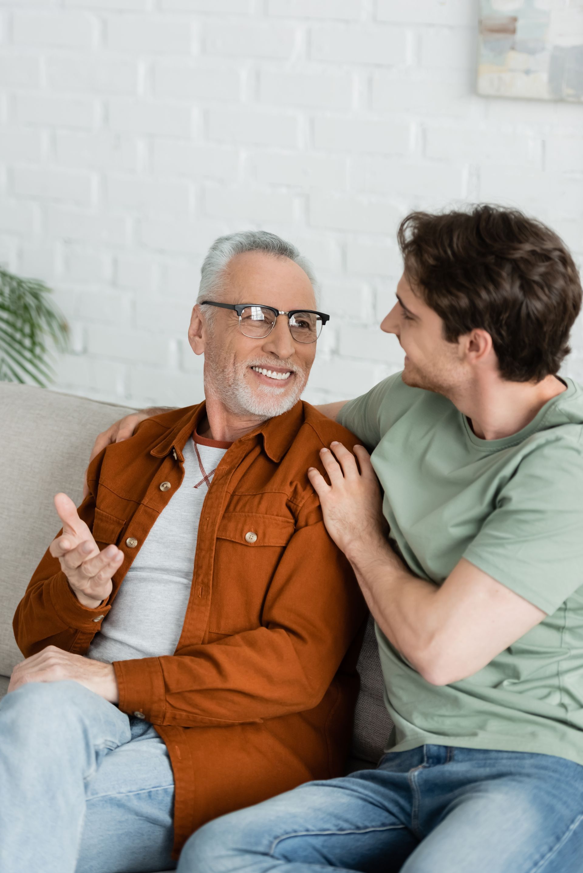 Man in orange shirt gestures, talking to another man with arm around him on a couch.