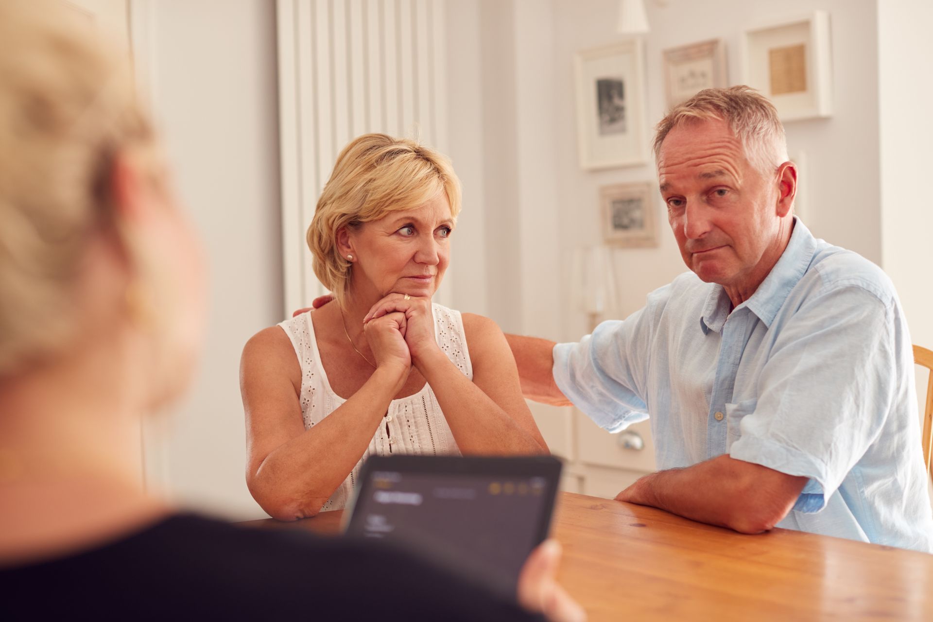 Couple at a table, listening to a person holding a tablet. Man has hand on woman's shoulder, all look concerned.