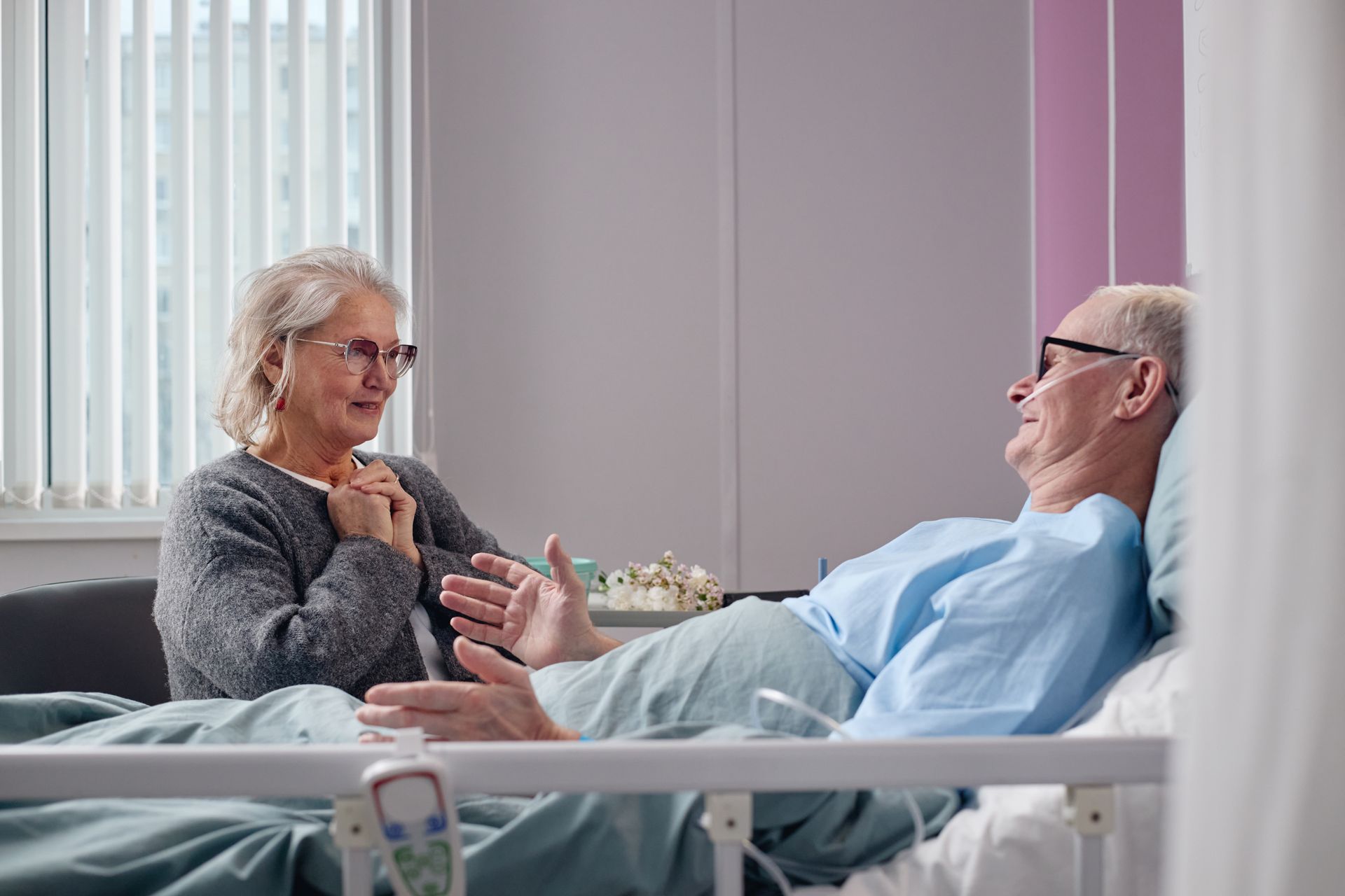 Woman sitting by a man in a hospital bed, talking with her hands.