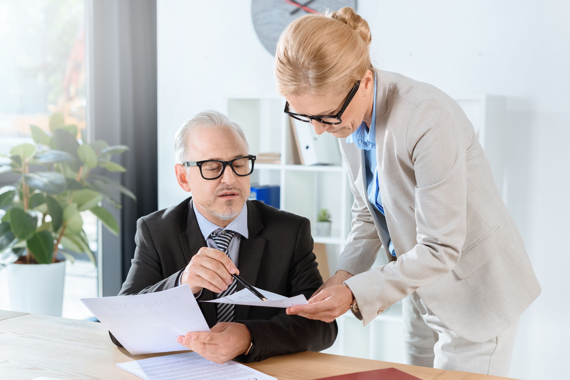 Two people reviewing documents in an office. One points to a paper while the other holds a pen, both wearing glasses.