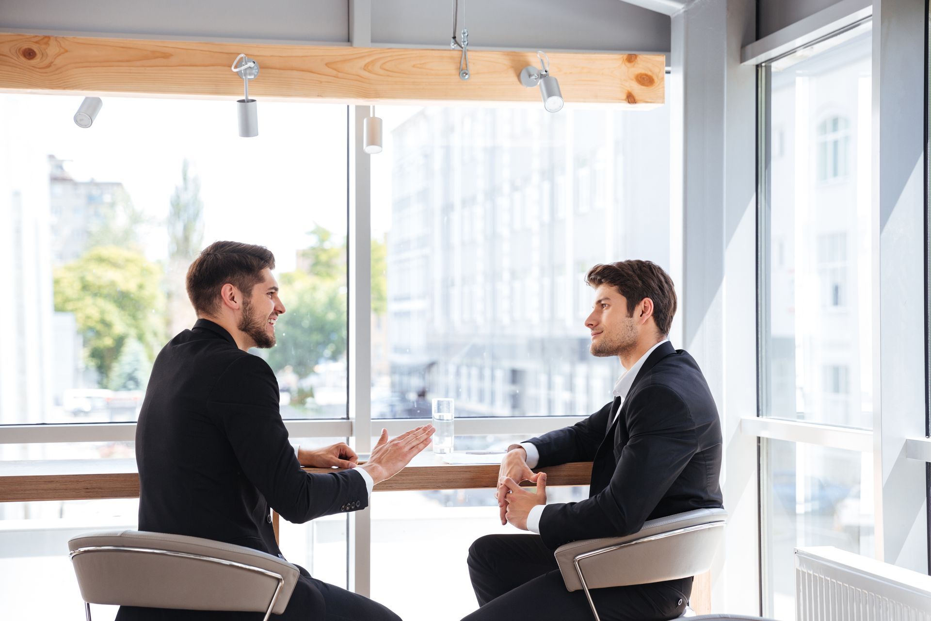 Man in orange shirt gestures, talking to another man with arm around him on a couch.