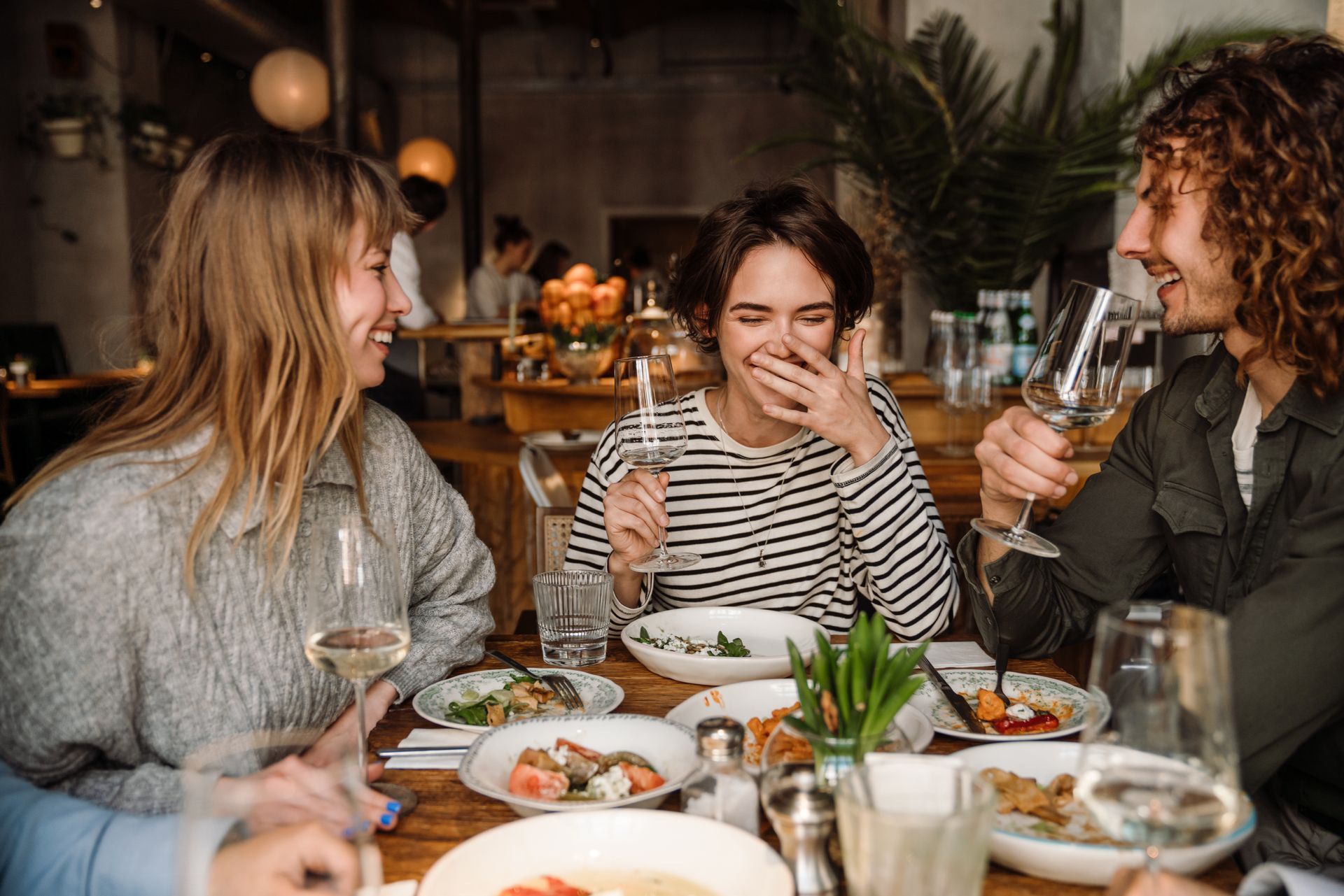 Three friends laugh while dining at a restaurant, enjoying food and wine.