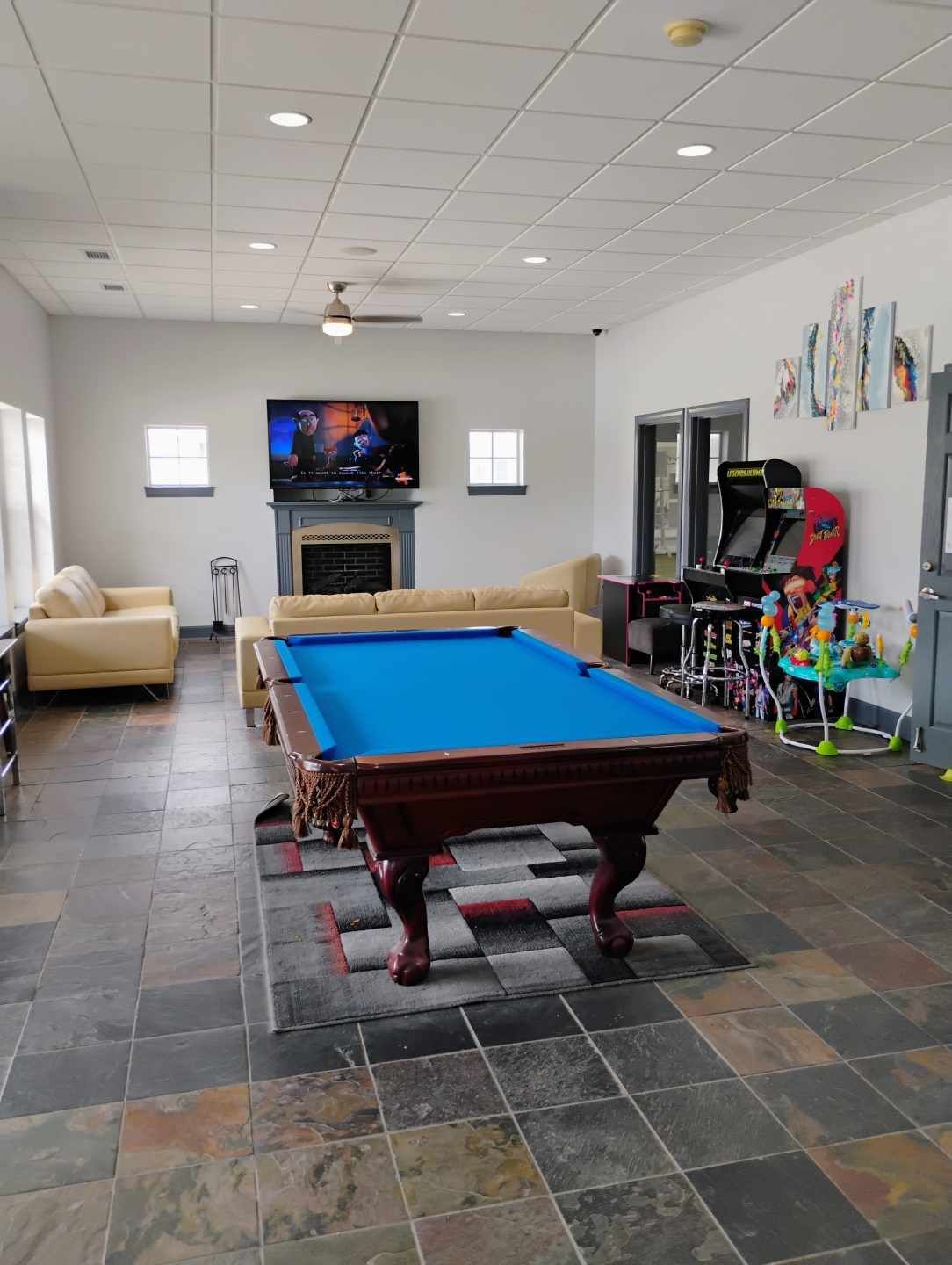 Pool table in a lounge area with couches, TV, fireplace, arcade games, and stone tile floor.