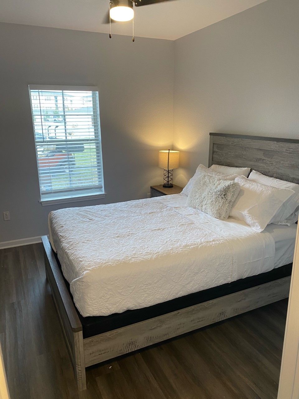 Bedroom with bed, lamp on nightstand, window with blinds, and wood-look flooring.
