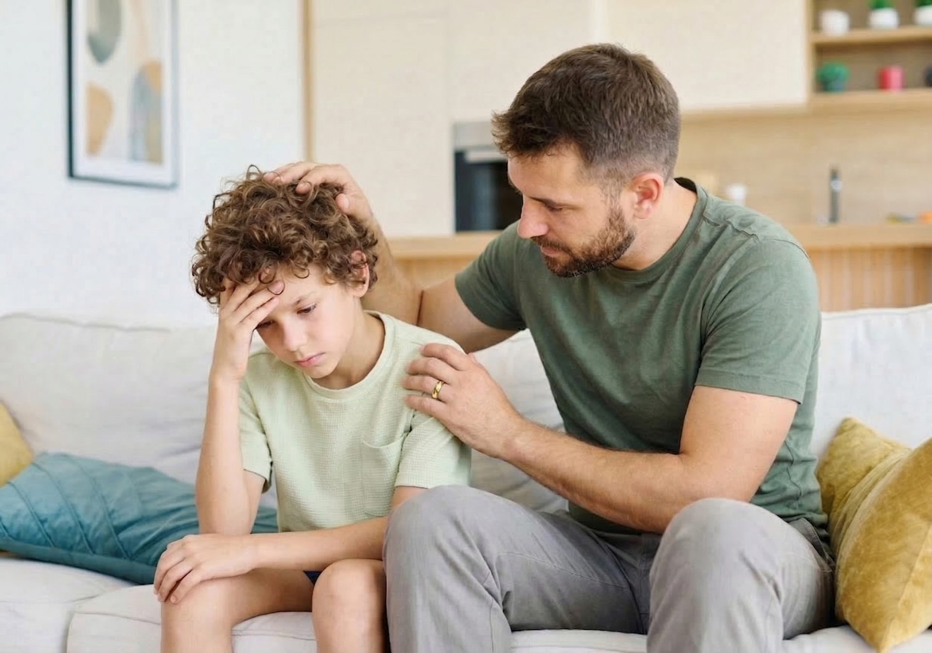 A parent sits on a couch, comforting a distressed child by placing a hand on their head and shoulder.