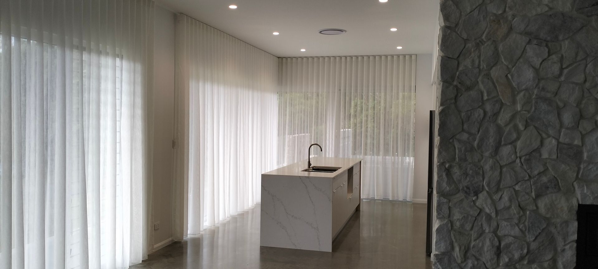 White Kitchen With Marble Island, Sheer Curtains, and Stone Wall — Seaview Blinds & Shutters in Woodbury, QLD