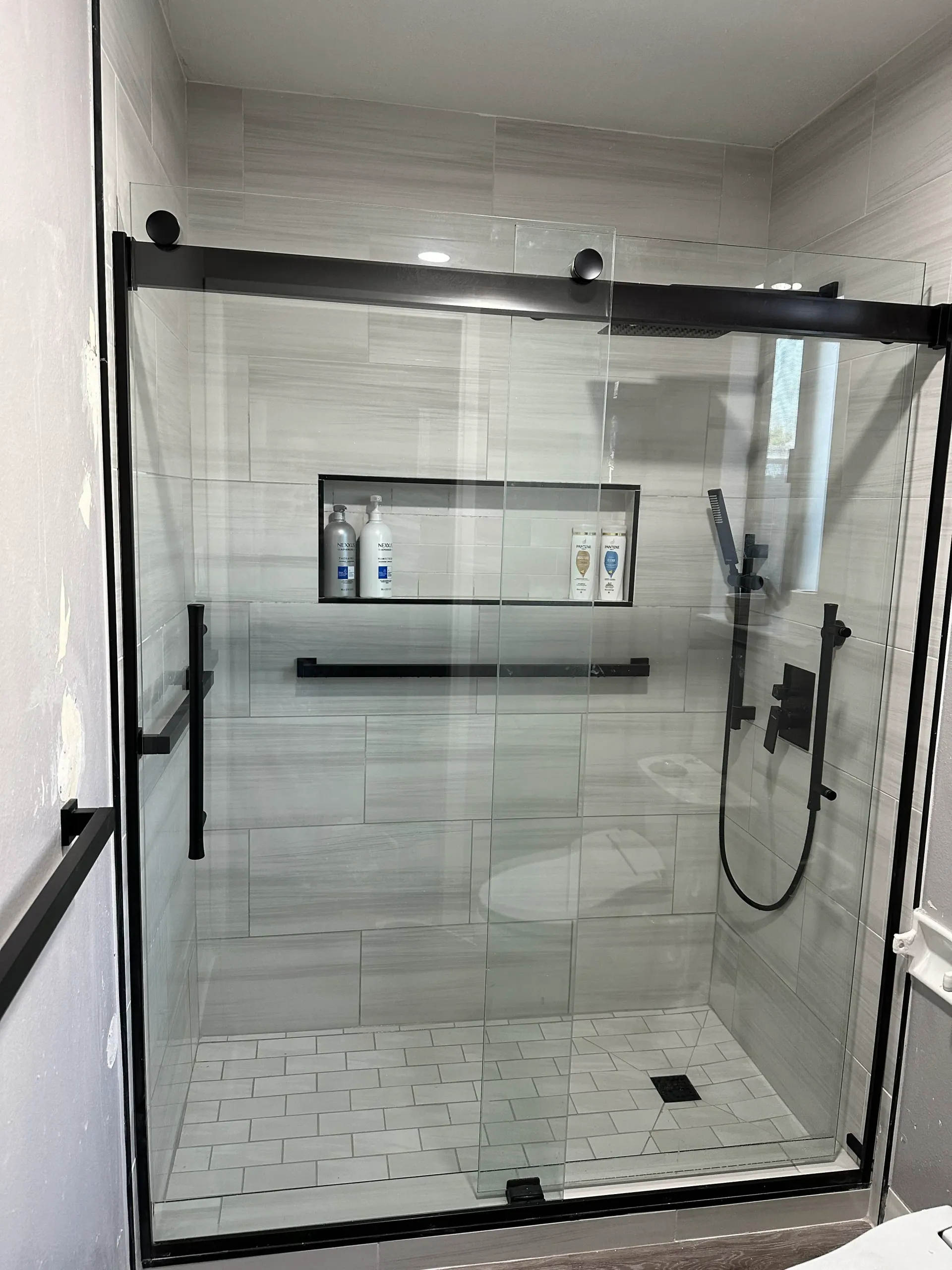 Black-framed glass shower with gray tile and black fixtures; a built-in shelf with toiletries.