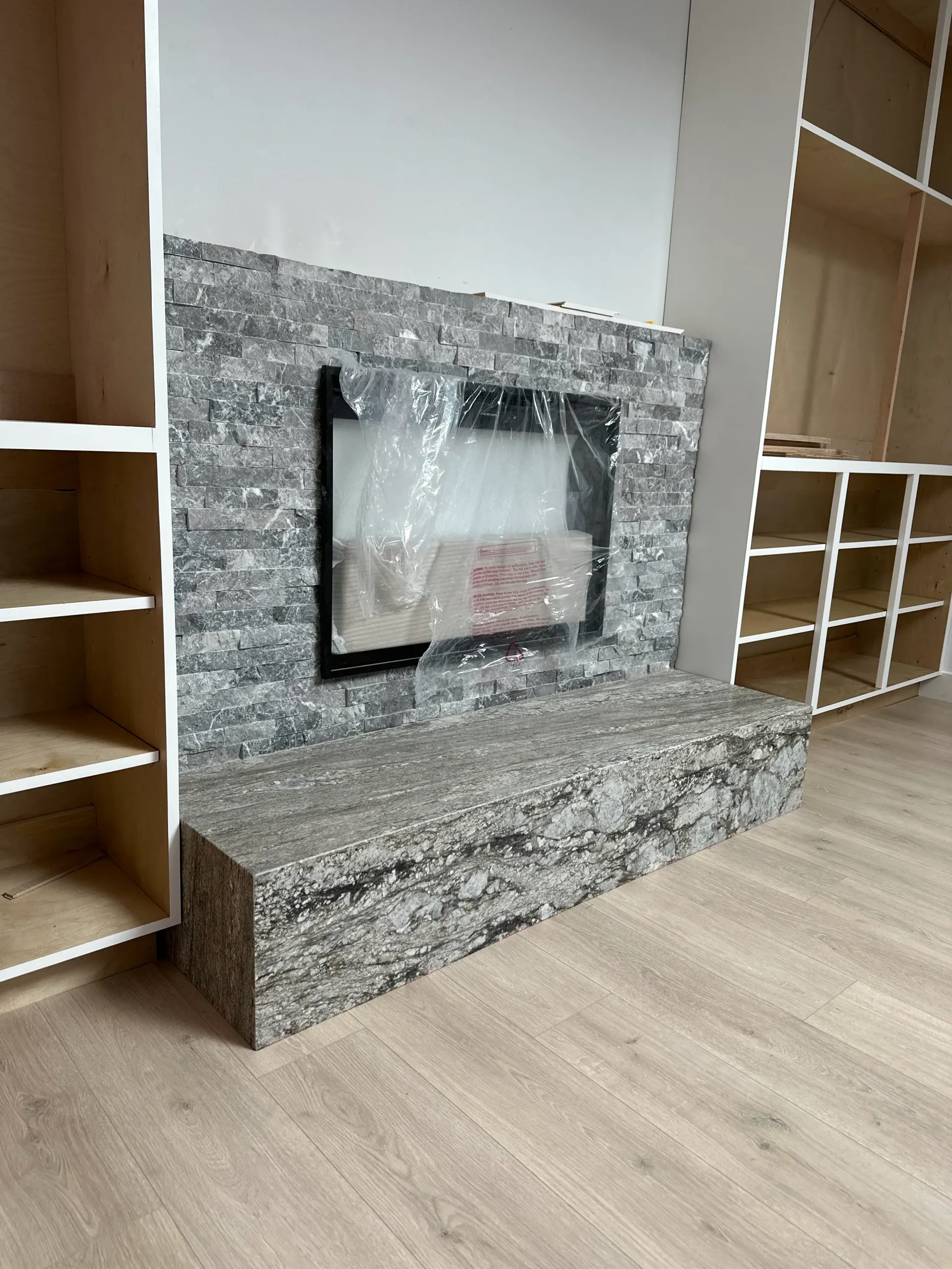 Fireplace with gray stone surround and bench, flanked by unfinished bookshelves on wood floor.
