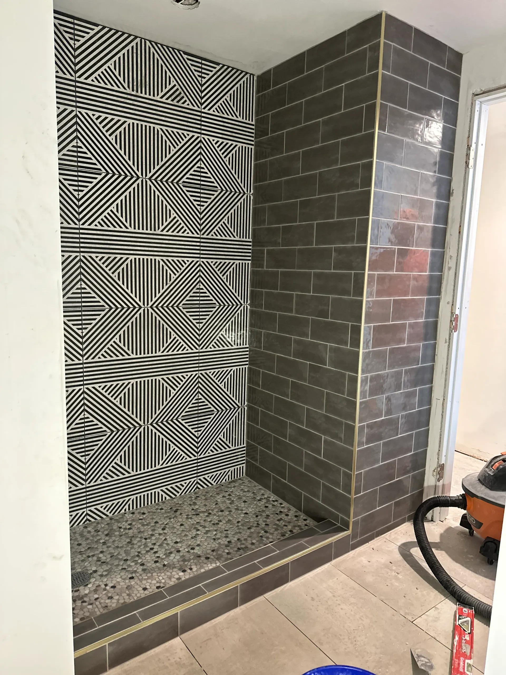 Shower with patterned black/white tile on the left wall, dark gray brick tile on the right, and a terrazzo-style floor.