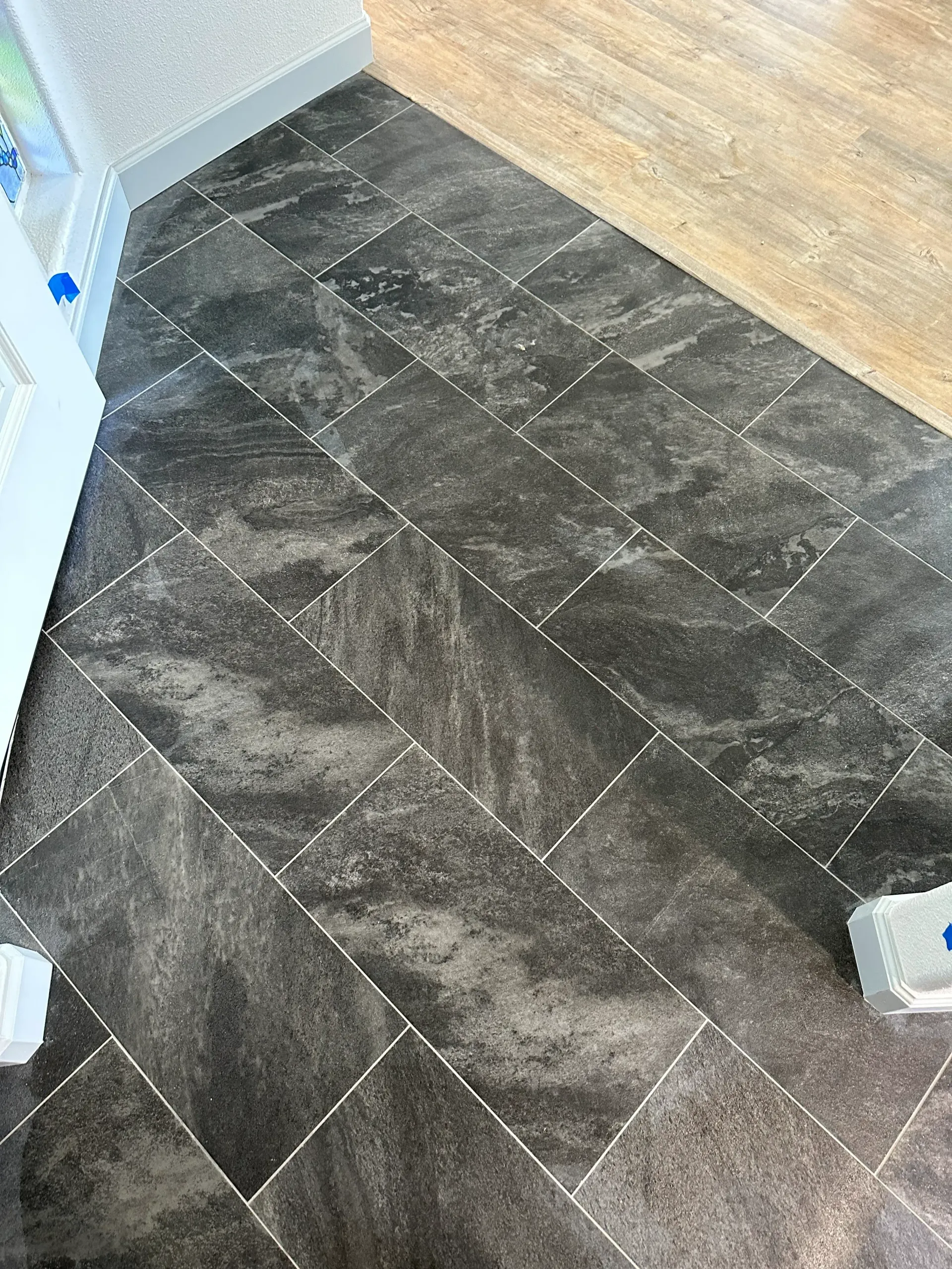 Dark gray patterned tile flooring laid in a herringbone pattern, next to light-colored wood floor and white trim.
