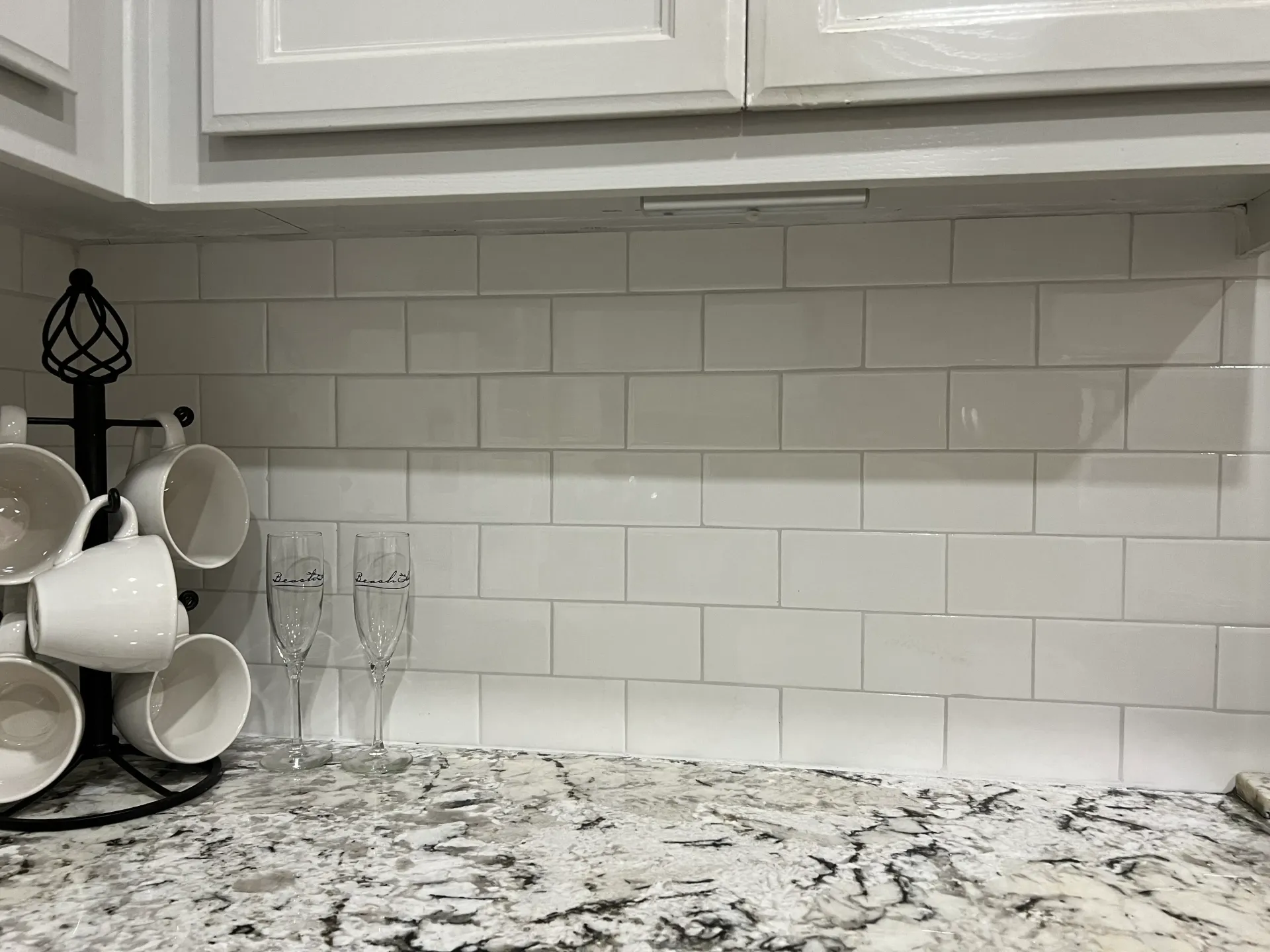 White kitchen with countertop, white tile backsplash, mugs, champagne flutes, and cabinets.