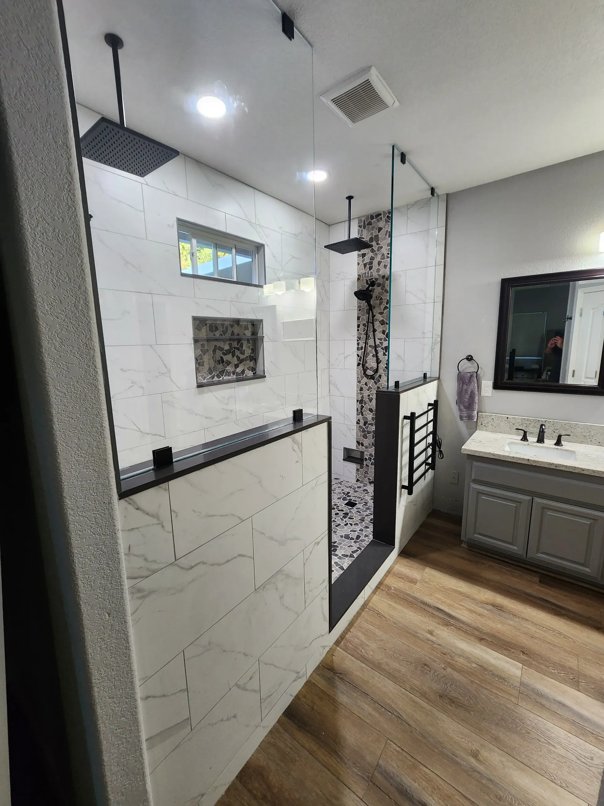 Modern bathroom with marble-look tile, glass shower, double vanity, and wood-look flooring.