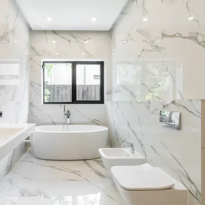 Bright white bathroom with marble tile, a tub, toilet, and window.
