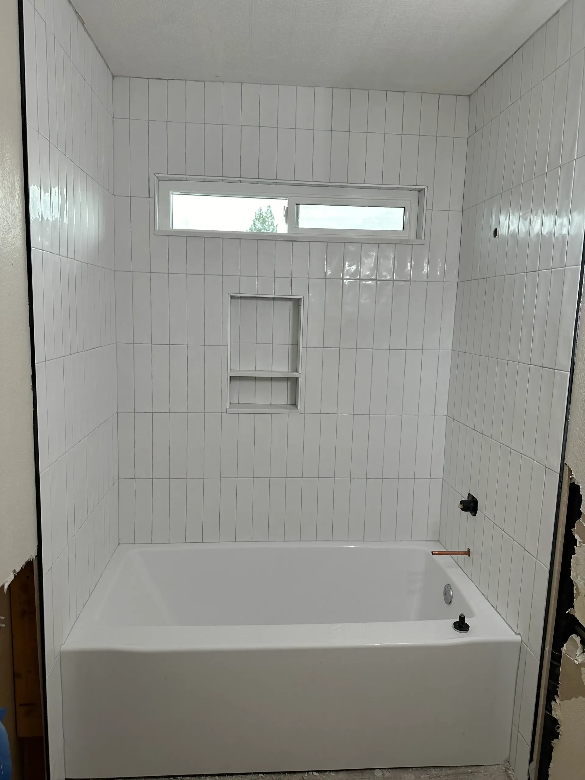 White tiled bathroom with a bathtub, window, and inset shelf.