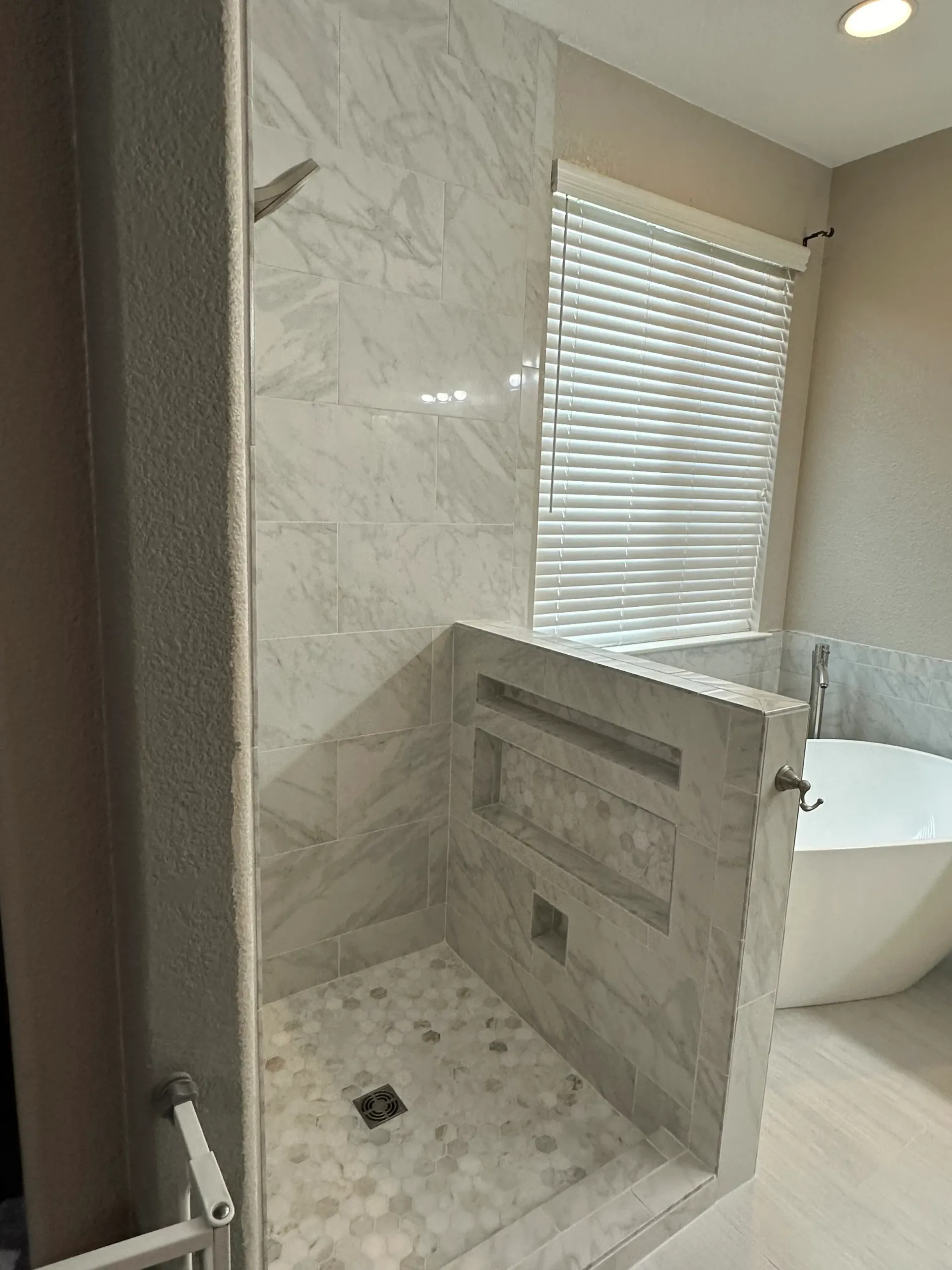 Modern bathroom with marble-tiled shower, built-in shelves, and a standalone tub near a window with blinds.
