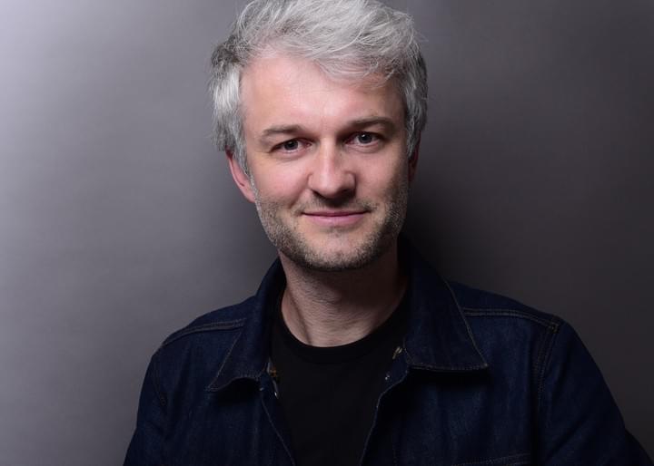 Ein Mann mit ergrauendem Haar lächelt vor einem grauen Hintergrund. Er trägt eine dunkle Jeansjacke und ein schwarzes Hemd.