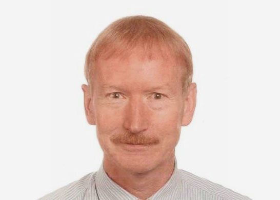 Ein Mann mit heller Haut, kurzem Haar und Schnurrbart blickt in einem Studio nach vorn.