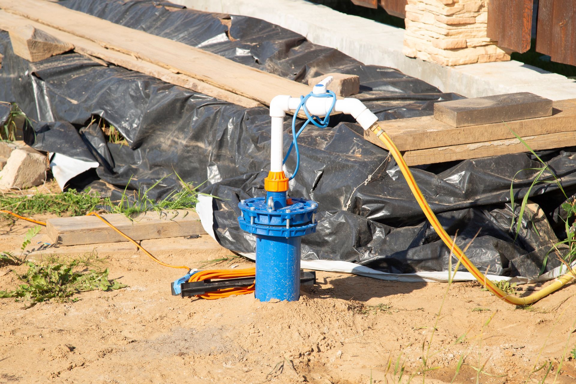 Blue water pump with yellow hose on sandy construction site, beside black tarp and stone steps
