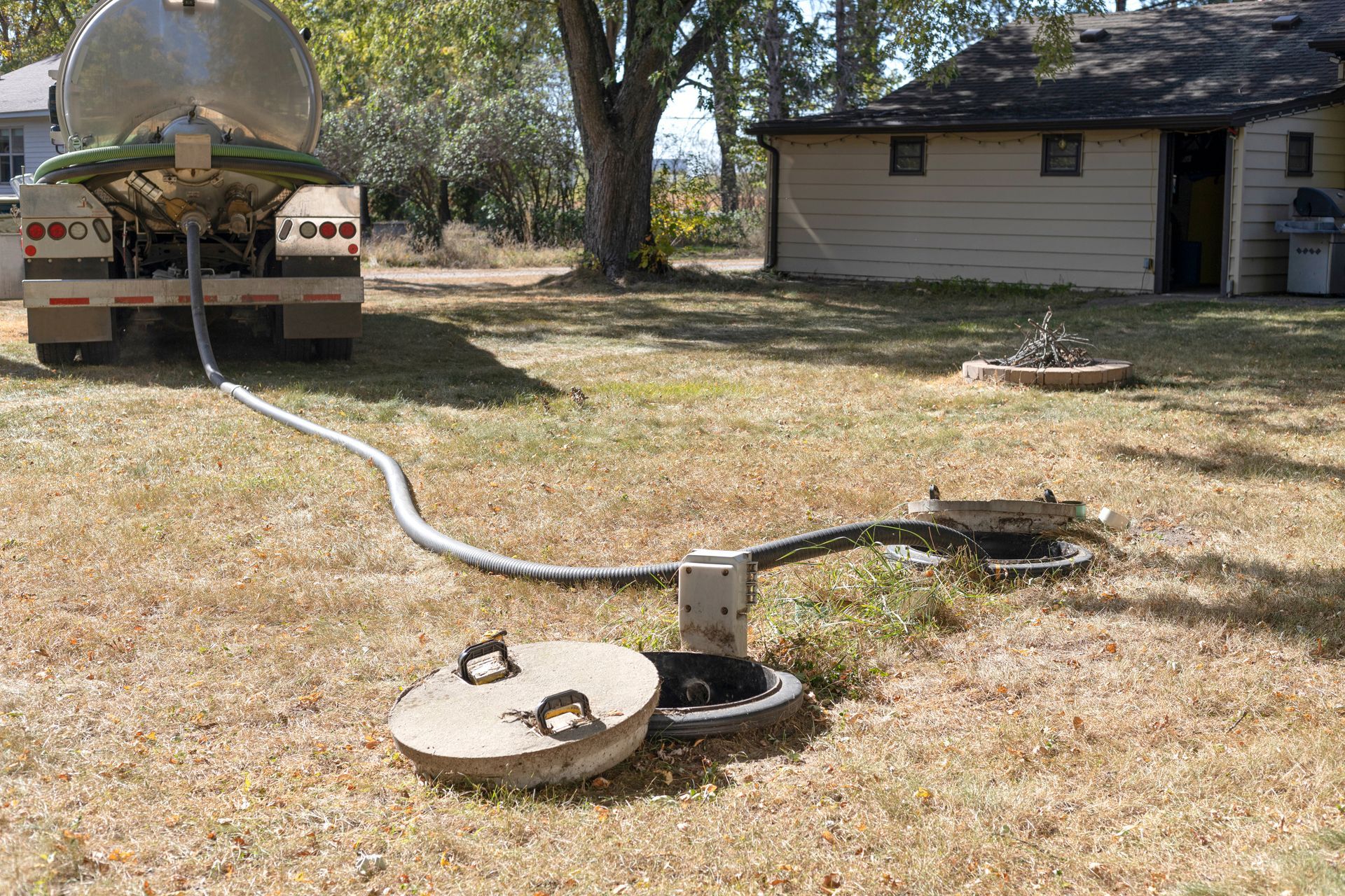 Backyard scene with a long hose or pipe connecting a tank to two open covers on dry grass.