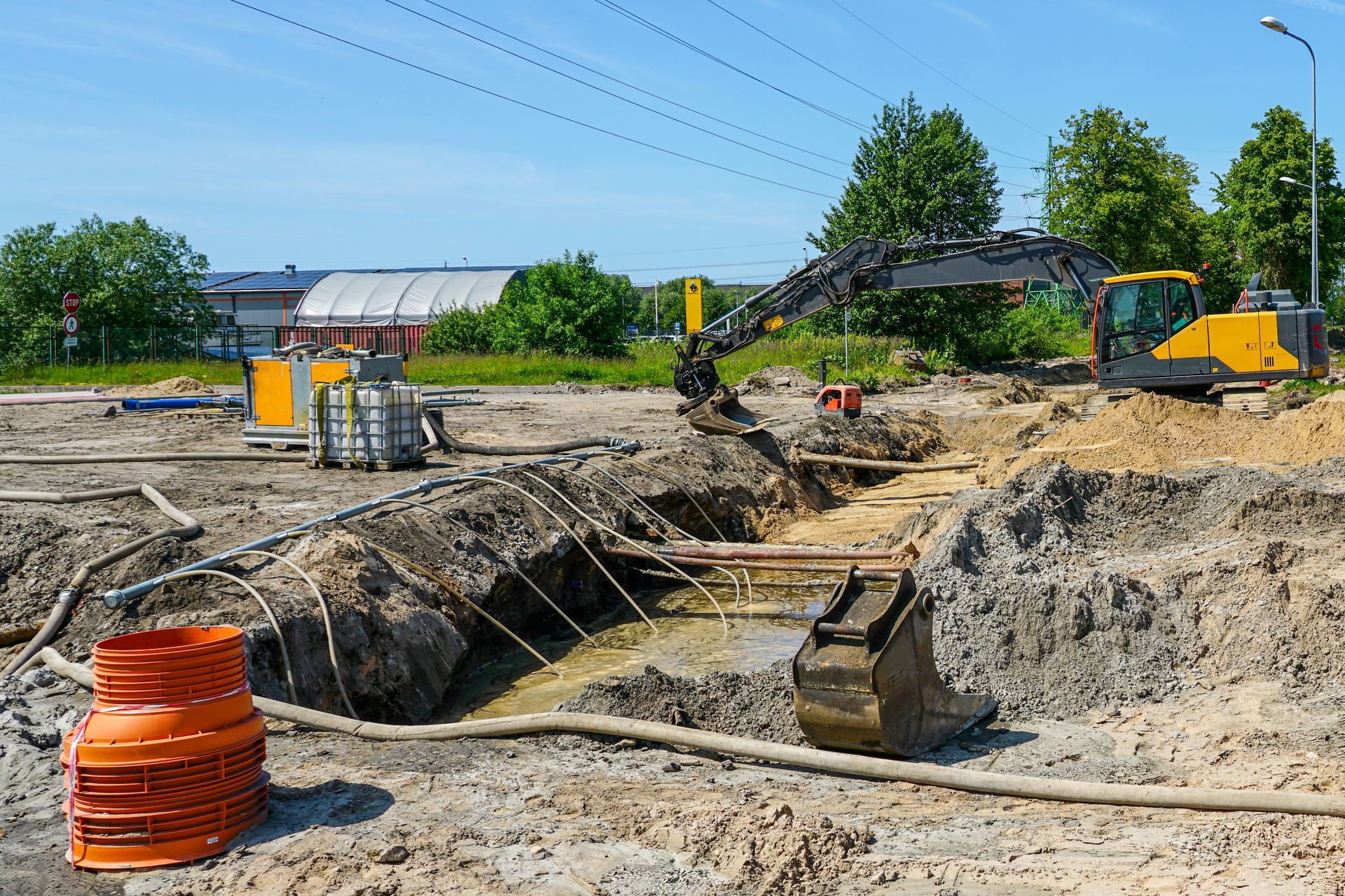 Construction site with excavator, trenches, and orange utility pipes under a blue sky