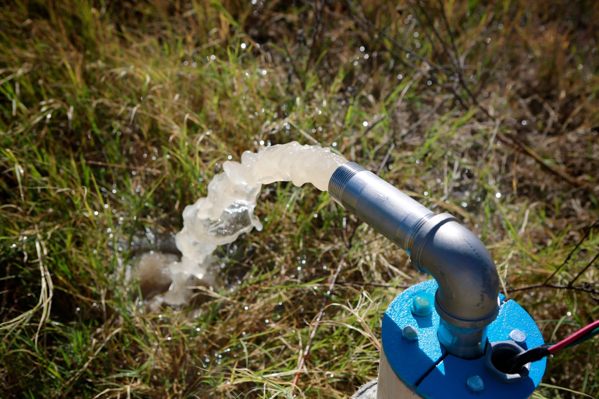 Water spraying from a metal pipe attached to a blue pump on grass outdoors