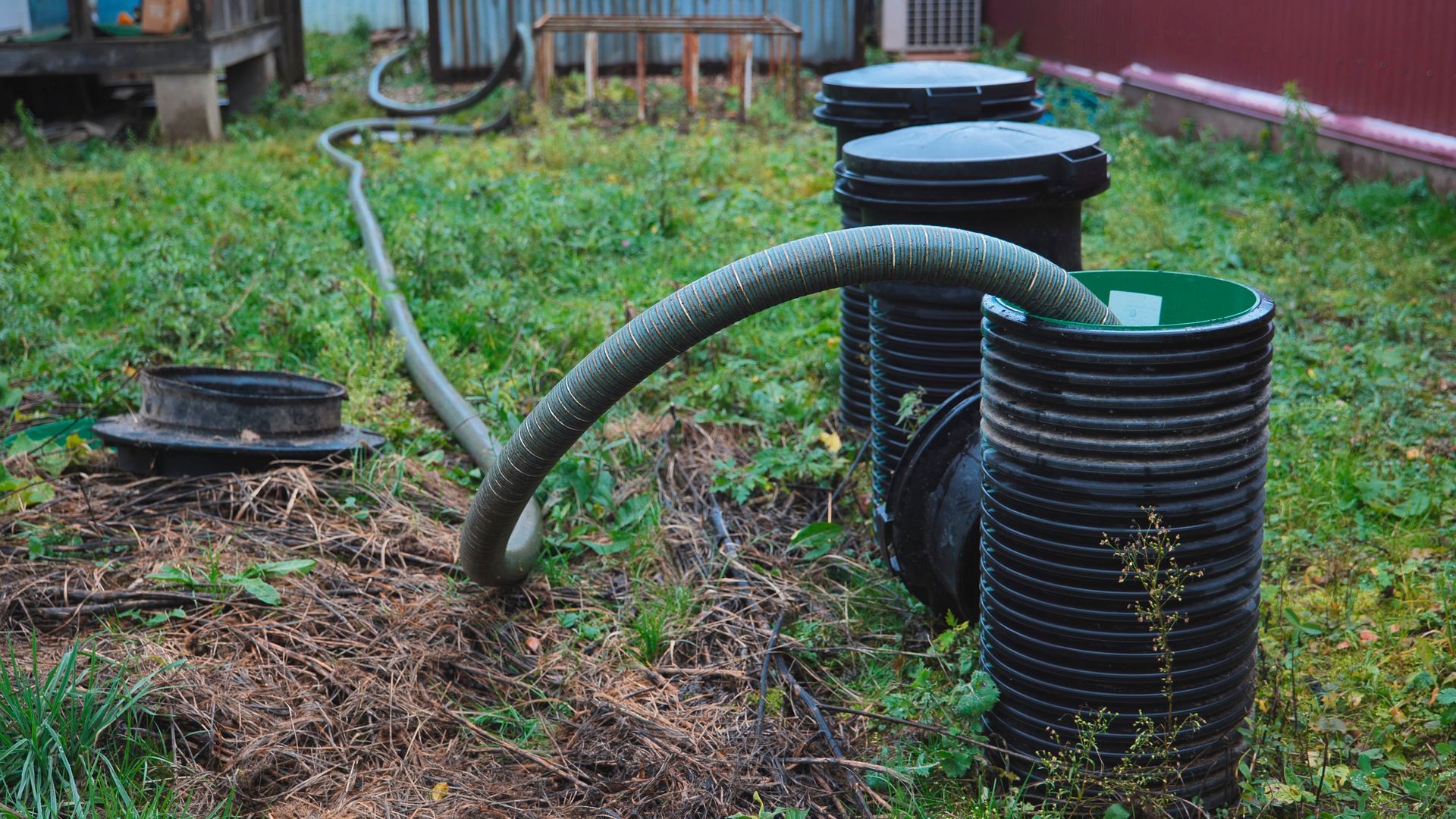 Black corrugated pipes and hoses arranged in a grassy yard near a house, suggesting drainage work