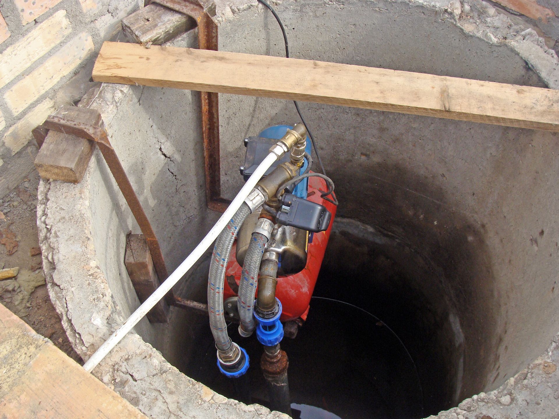 Open manhole with red pump and pipes inside a concrete well, topped by a wooden plank.