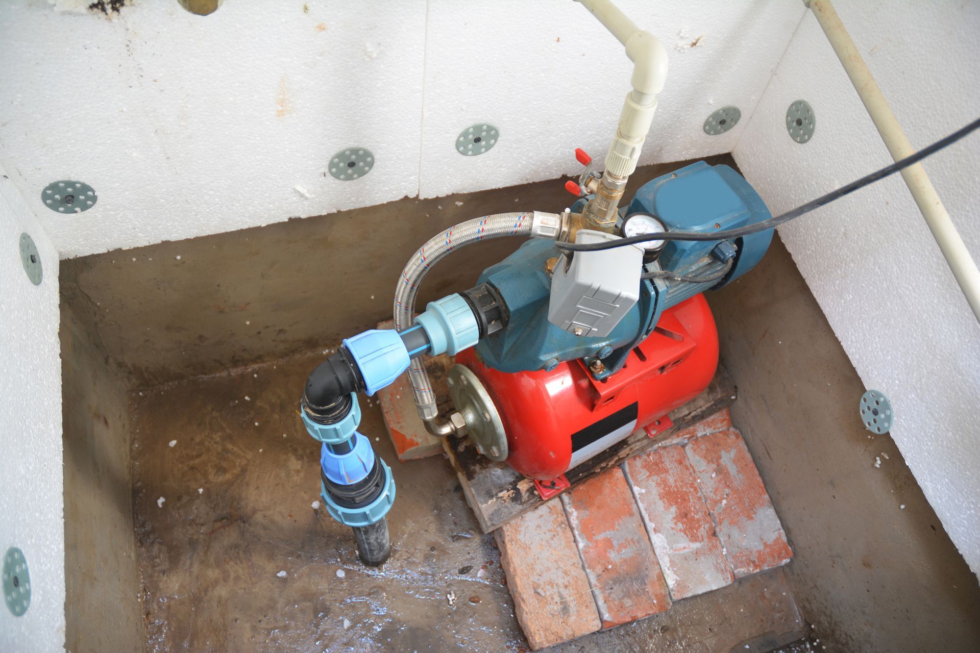 Red water pump and blue pipes installed in a small utility pit with brick floor and concrete walls